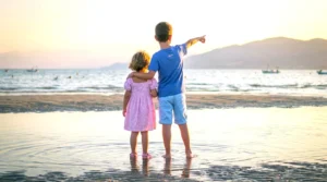 Deux enfants au bord de la mer, l’un en robe rose et l’autre en tenue bleue, regardant ensemble l’horizon au coucher du soleil, avec des bateaux et des montagnes en arrière-plan.