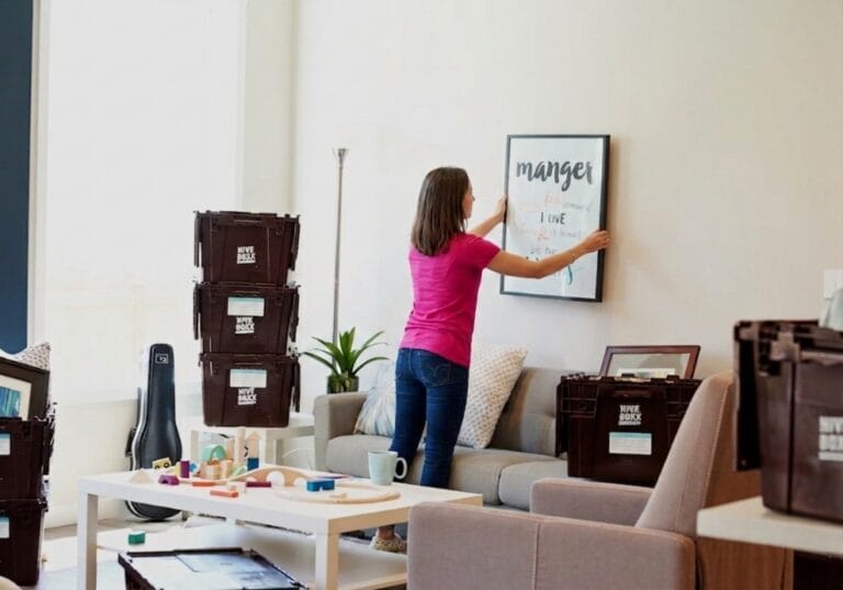 Personne accrochant un cadre sur le mur d’un salon en cours d’aménagement, entouré de cartons de déménagement, d’un canapé gris, d’une table basse avec jouets colorés et d’un étui de guitare.