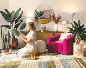 Entretien plantes d'intérieur dans un salon moderne avec fauteuil magenta et miroir rond. Une personne s'agenouille pour sauver une plante qui meurt en ajustant son cycle d'arrosage. Le décor inclut un terreau de rempotage, un engrais naturel posé sur une table, et une luminosité douce qui éclaire les grandes plantes vertes.