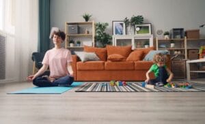 Salon moderne baigné de lumière naturelle, une maman médite sur un tapis de yoga bleu tandis qu’un enfant joue avec des blocs colorés sur un tapis rayé, créant une atmosphère paisible et familiale.